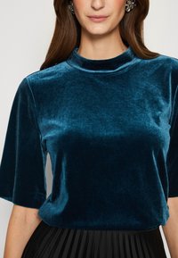 Tyrkysová sametová blůza s krátkými rukávy a vysokým výstřihem. Látka má jemnou, lesklou textura, doplněná černou plisovanou sukní.