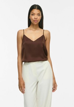 Kvinde står iført en brun ærmeløs camisole-top med tynde stropper og et par hvide bukser med høj talje, vendt fremad mod en ensfarvet baggrund.