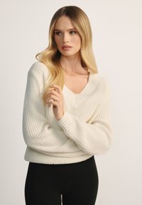 Kobieta z długimi blond włosami, ubrana w biały dziergany sweter z dekoltem w serek i czarne spodnie, patrząca w bok z założonymi ramionami.