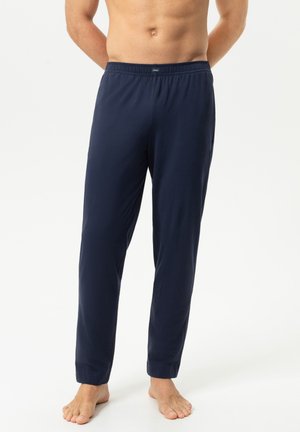 Man die donkerblauwe, loszittende pyjamabroek draagt, blootsvoets voor een witte achtergrond staat met handen achter de rug en zichtbare blote torso.