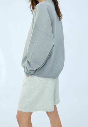 Henkilö, jolla on päällään ylisuurikokoinen harmaa collegepaita ja vaaleanharmaat polvipituiseen ulottuvat shortsit, seisoo yksinkertaisen vaalean taustan edessä.