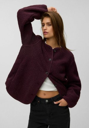 Femme aux longs cheveux bruns portant une veste polaire violet foncé boutonnée, un top court blanc et un jean noir, posant avec un bras levé.