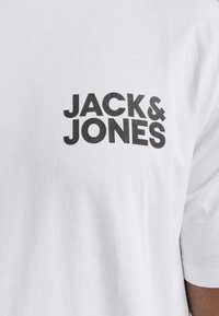 Biely bavlnený tričko s čierne potlačeným logom s textom "JACK & JONES" na ľavej hornej časti hrudi. Krátke rukávy a hladká textura.