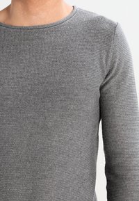 Pull gris à manches longues avec un col rond. Texture tricotée avec un motif subtil. Le tissu semble doux et léger.