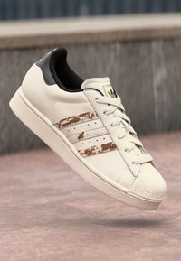 White leather sneaker with three faded brown stripes, textured toe cap, black collar, and light-colored laces. Gold logo on tongue.