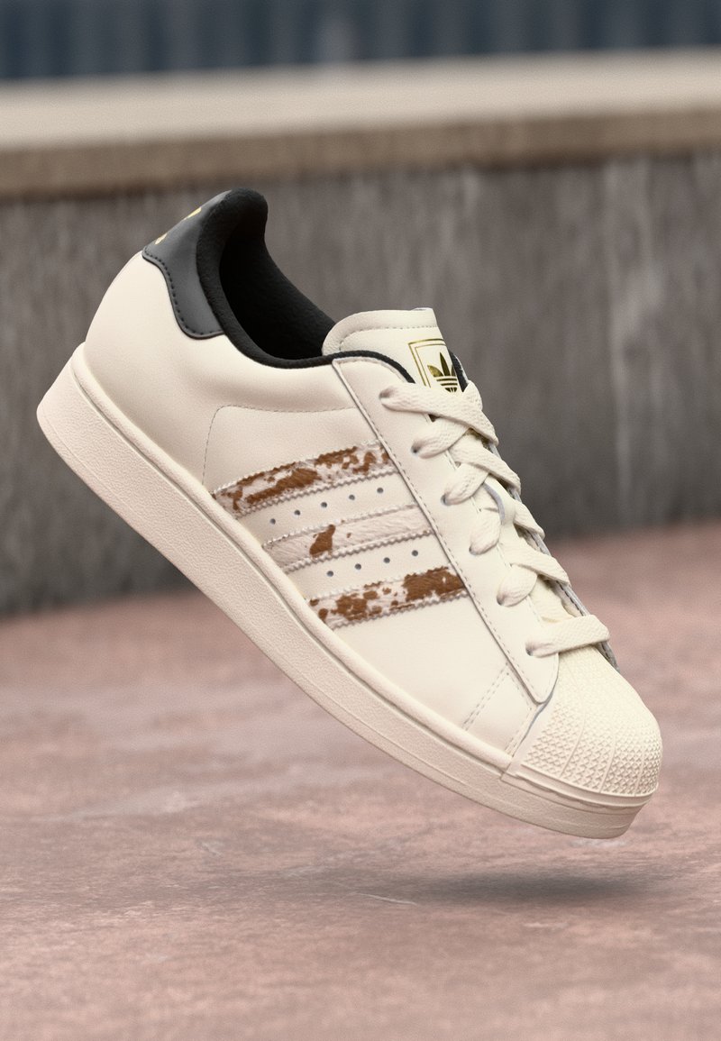 White leather sneaker with three faded brown stripes, textured toe cap, black collar, and light-colored laces. Gold logo on tongue.