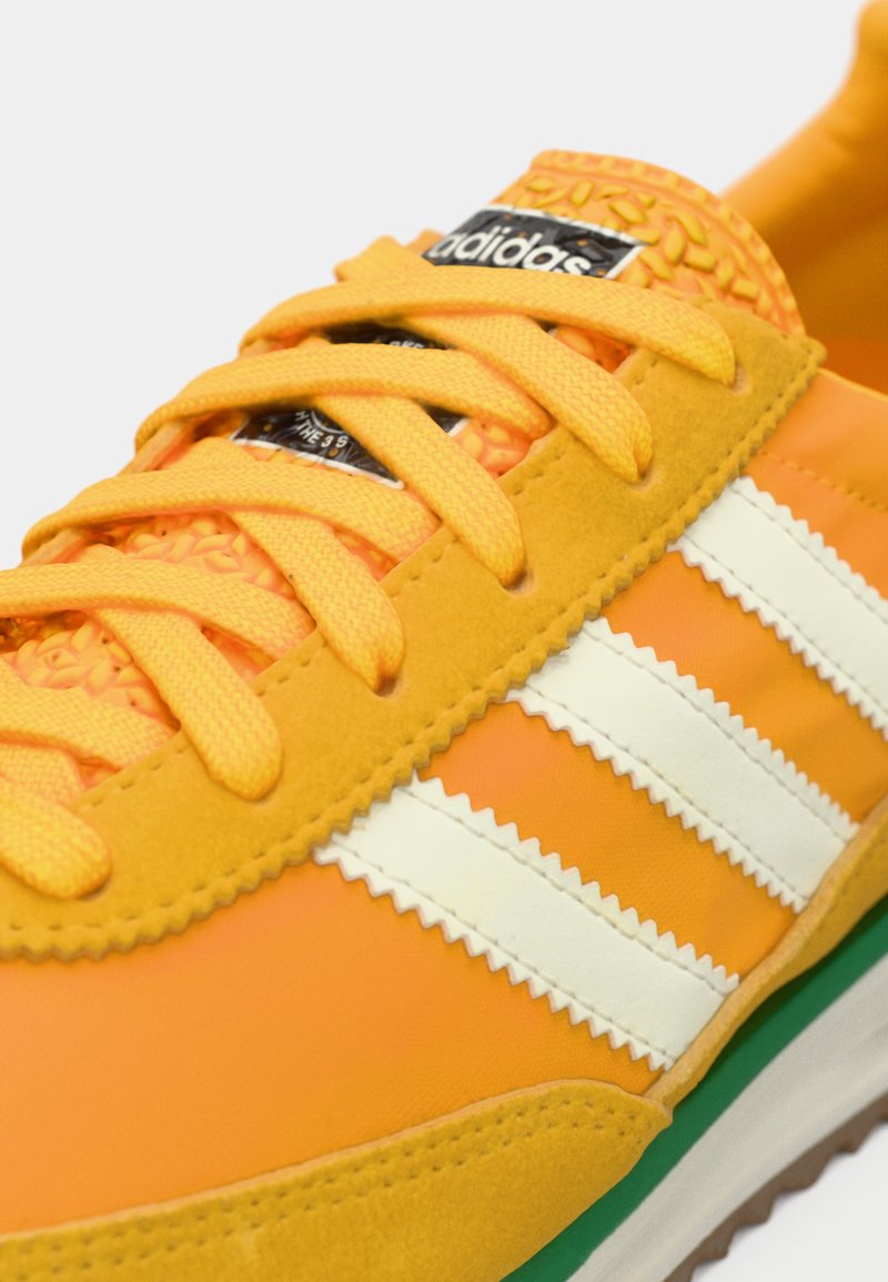 Zapatilla Adidas de color naranja brillante con cordones amarillos, tres franjas blancas y lengua texturizada sobre un fondo blanco.