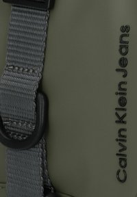 Borsa Calvin Klein Jeans olive green in tessuto strutturato. Presenta una tracolla grigia e dettagli in metallo nero con logo in rilievo.