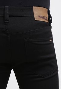 Jeans negros con una textura suave, que cuentan con un parche de cuero marrón y costuras en contraste en los bolsillos traseros. Logo grabado en el parche.
