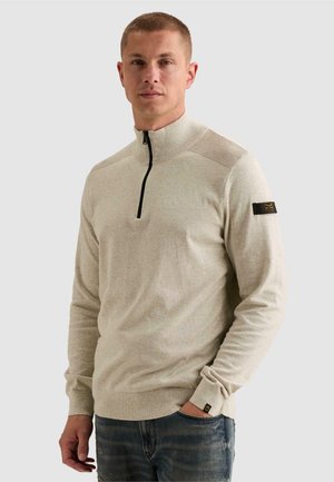 Homme aux cheveux courts portant un pull beige à manches longues avec une fermeture éclair partielle et un jean bleu foncé, debout avec une main dans la poche.