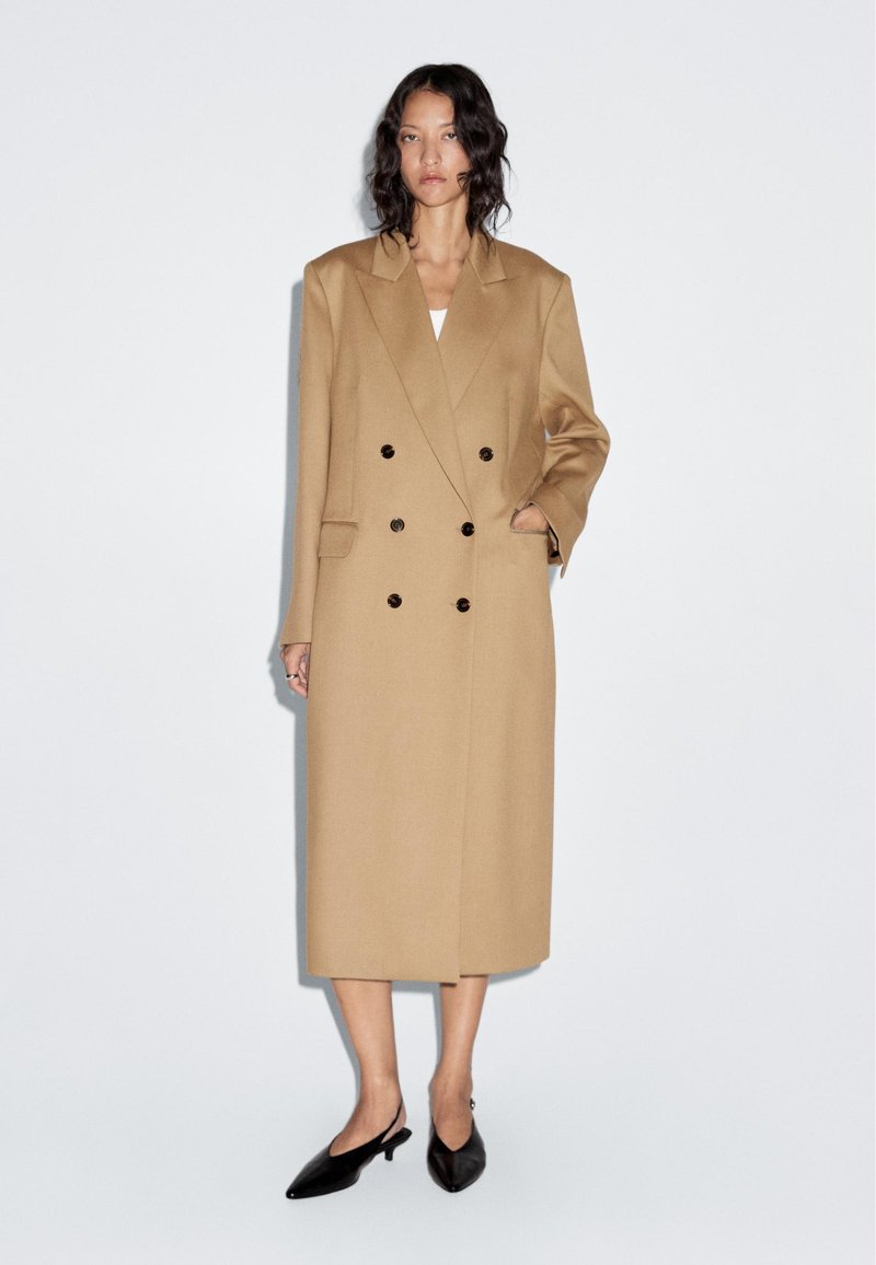 Beige double-breasted coat with large lapels, side pockets, and black buttons; knee-length with a smooth texture, worn over a white top.