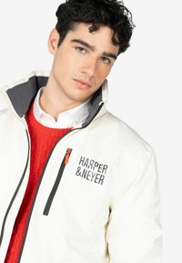 Chaqueta blanca con cuello gris, con el texto negro "HARPER & NEYER" y un distintivo tirador de cremallera naranja. Suéter rojo por debajo.