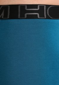 Tyrkysová elastická tkanina vesty nebo tílka s širokým černým elastickým pásem, na jehož vrchu je stříbrné logo. Hladký povrch, přiléhavý střih.