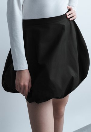 Personne portant un haut blanc à manches longues et une mini-jupe noire style ballon, tenant l'ourlet de la jupe d'une main.