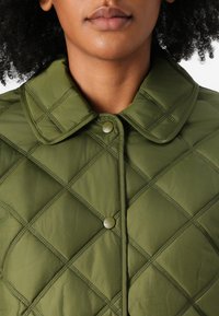Veste matelassée vert olive avec un col pointu, dotée d'un cousu en diamant et d'une fermeture à boutons-pression, fabriquée à partir d'un matériau lisse.