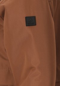 Bruine jas gemaakt van gladde stof, met een zwart logo-opdruk op de mouw. Het ontwerp is elegant met minimale stikdetails.
