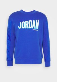 Blå sweatshirt i mjukt tyg, med ordet "JORDAN" i djärva vita bokstäver. Har glänsande blå detaljer på ärmarna.
