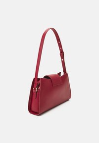 Červená kožená kabelka s úzkým, strukturovaným designem, nastavitelným ramenním popruhem a klopovým uzávěrem. Hladká textura a minimalistický styl.