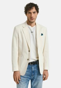 Blazer texturé blanc cassé avec des revers en pointe, deux boutons et poches latérales, associé à un t-shirt blanc et un jean bleu clair.