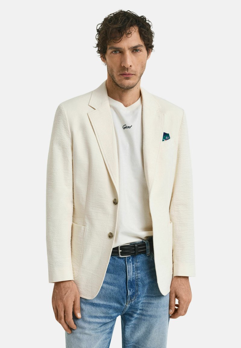 Blazer texturé blanc cassé avec des revers en pointe, deux boutons et poches latérales, associé à un t-shirt blanc et un jean bleu clair.