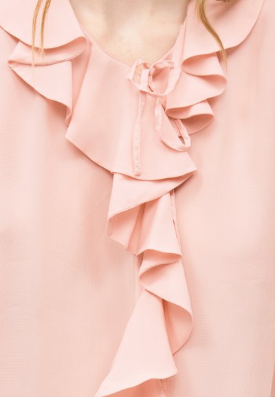 Gros plan sur un chemisier rose pâle avec des volants superposés et un nœud noué au niveau de l'encolure.