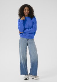 Sweat-shirt bleu avec des poignets et un ourlet côtelés, associé à un jean ample bleu clair et des baskets grises. Le mannequin est en pose décontractée.