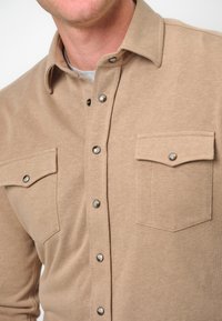Camicia button-up beige in tessuto morbido, con colletto a punta, due tasche sul petto e bottoni a pressione metallici lungo la patta anteriore.