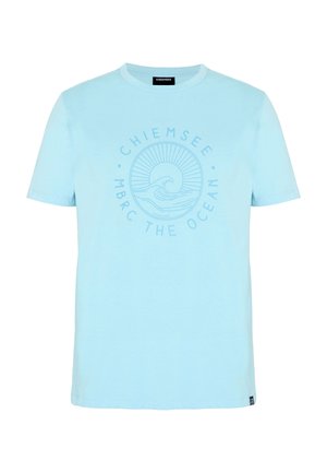 Junger Mann in hellblauem T-Shirt und Shorts mit ozeanbezogenen Logos, steht entspannt mit einer Hand in der Tasche und der anderen in der Nähe seines Halses.
