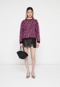 Maglione con stampa leopardata rosa e nera abbinato a una mini gonna di pelle nera. Tiene in mano una borsa nera dal design strutturato e indossa scarpe a punta color nude.