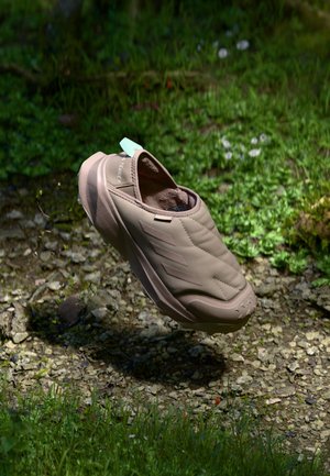 Beige slip-on sneaker with a textured, padded upper, featuring side accents and a rugged sole, positioned above uneven ground with grass and stones.