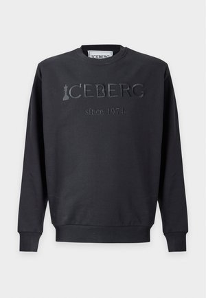 Young man wearing a black sweatshirt with "ICEBERG since 1971" text, dark jeans, and white sneakers, standing against a plain white background.