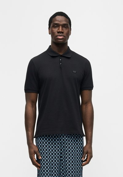 Άνδρας που φοράει μαύρο πόλο με κοντά μανίκια και μπλε παντελόνι με σχέδια, στέκεται μπροστά σε λευκό, απλό φόντο, κοιτάζοντας εμπρός.