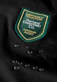 Černá tkanina s vyšívanou nášivkou v zelené a zlaté barvě s nápisem "Portland Outerwear Co." Na povrchu jsou viditelné kapky vody.