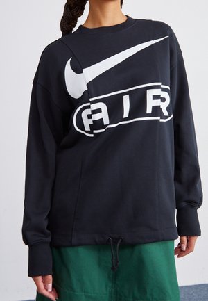 Osoba nosící černou mikinu Nike s velkým bílým logem a zelenou sukní stojící na neutrálním pozadí.