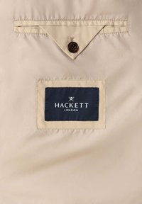 Tissu beige avec une étiquette rectangulaire Hackett London cousue sous un rabat triangulaire maintenu par un bouton marron.