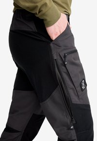 Person som bär svarta och mörkgrå cargobyxor med dragkedje- och klafffickor, som stoppar en hand i en sidoficka, klädd i en olivgrön topp.