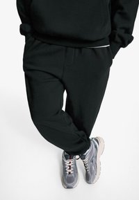 Pantalons de survêtement noirs à coupe décontractée, dotés d'une taille élastique et de poches latérales, associés à des baskets grises avec plusieurs textures et couleurs.