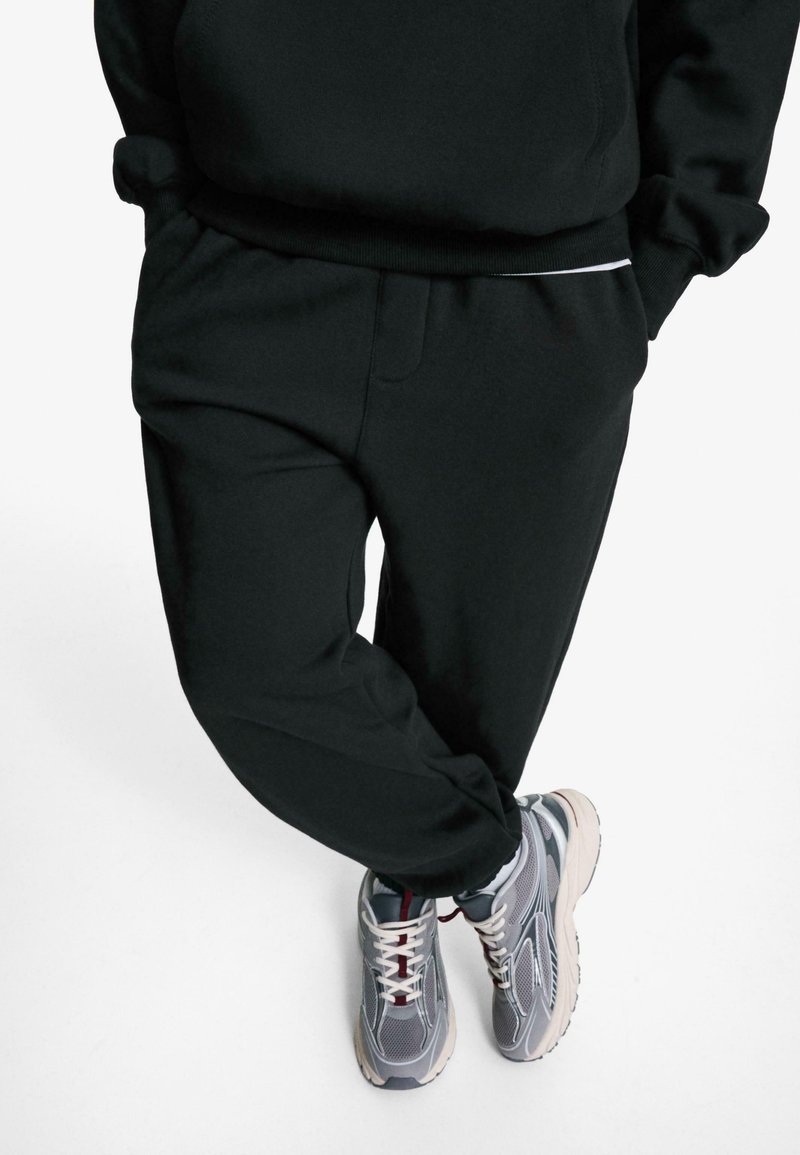 Pantalons de survêtement noirs à coupe décontractée, dotés d'une taille élastique et de poches latérales, associés à des baskets grises avec plusieurs textures et couleurs.