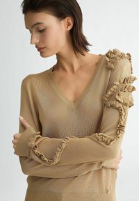Maglione beige a scollo a V con delicati dettagli arricciati sulla spalla sinistra e sui polsini. Tessuto leggero e strutturato, design aderente.