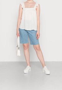 Top bianco senza maniche con spalline arricciate e merletto, abbinato a pantaloncini in denim azzurri. La modella tiene una piccola borsa bianca e indossa sneakers bianche.