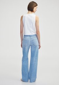 Jeans de mezclilla de tono azul claro, de campana y con cintura alta, decorados con patrones de tachuelas blancas, combinados con una blusa sin mangas blanca.