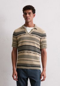 Koszulka polo z krótkim rękawem w jasnym beżu z czarnymi poziomymi paskami i wzorami. Posiada dekolt w serek i teksturowany materiał.
