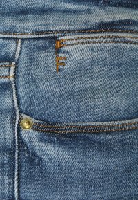 Tejido de mezclilla azul, que presenta un pequeño bolsillo trasero con un botón dorado, costuras naranjas y una sutil textura desgastada cerca del área del bolsillo.