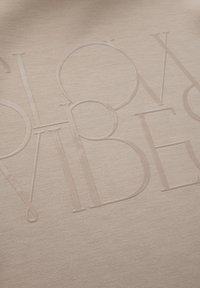 Beige textil med en texturerad yta där präglade bokstäver står "SLOW MOVIDES." Slät yta med en subtil lyster och mjukt tyg.