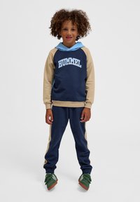 Marineblauwe en tan hoodie met capuchon, met een blauw "HUMMEL" logo; gecombineerd met bijpassende marineblauwe joggers en groene sneakers.
