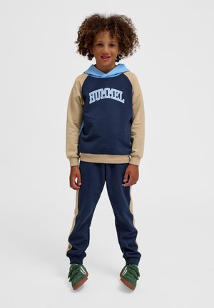 Granatowa i beżowa bluza z kapturem, z niebieskim logo "HUMMEL"; zestawiona z pasującymi granatowymi dresami i zielonymi sneakersami.