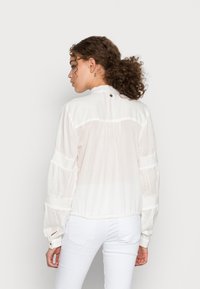 Blusa branca, leve, com gola alta, mangas longas com volume e punhos elásticos. Inclui um detalhe de botão nas costas e uma textura subtil.