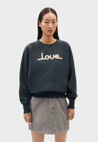 Felpa blu scuro con polsini e orlo a costine, caratterizzata da una grafica "love" color crema sulla parte anteriore. Indossata con una gonna in corduroy grigia.