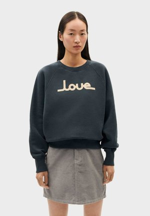 Felpa blu scuro con polsini e orlo a costine, caratterizzata da una grafica "love" color crema sulla parte anteriore. Indossata con una gonna in corduroy grigia.