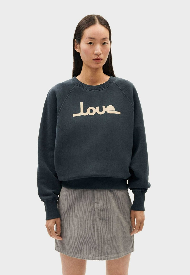 Felpa blu scuro con polsini e orlo a costine, caratterizzata da una grafica "love" color crema sulla parte anteriore. Indossata con una gonna in corduroy grigia.